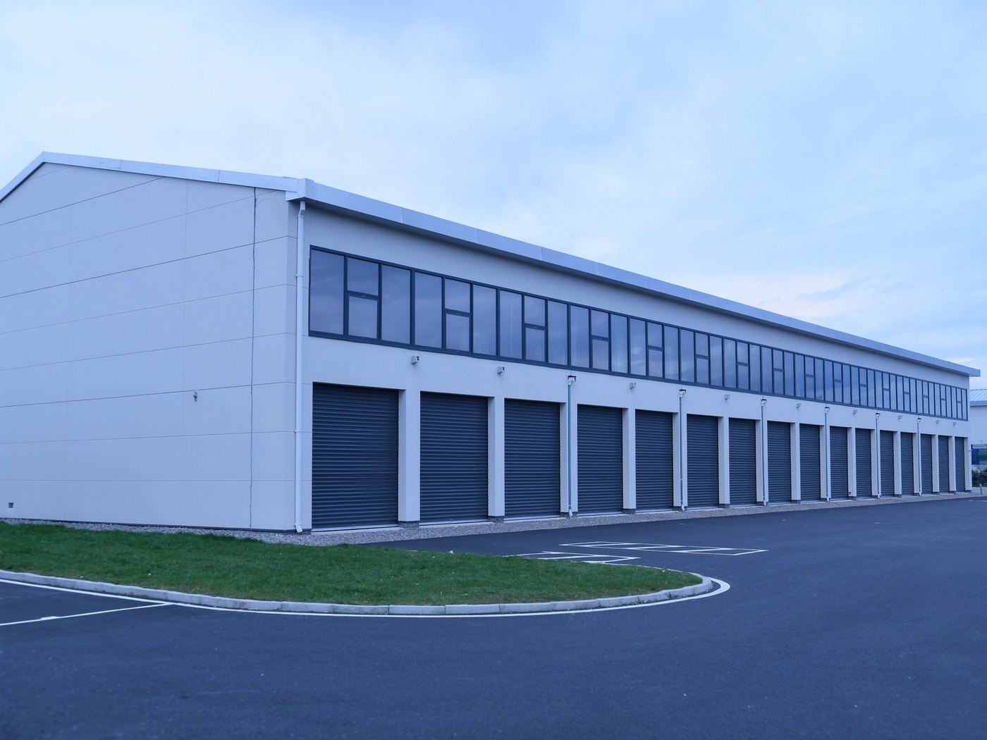 Vue d’ensemble d’un centre de self-stockage achevé : longue façade en bardage métallique clair, rangée de portes rideaux en acier fermées, bandeau vitré en hauteur, parking asphalté au premier plan
